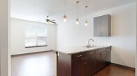 This single-floor unit offers a thoughtfully designed open-concept layout that seamlessly connects the kitchen and living area. The kitchen serves as the focal point, anchored by a peninsula with a granite countertop, an undermount double sink, and rich dark-wood cabinetry below. Above, sleek gray upper cabinets provide ample storage, while a trio of pendant lights and an adjustable track lighting system illuminate the space with style. Stainless steel and black appliances — including a refrigerator, range, and built-in microwave — complete the well-equipped kitchen. The living area extends naturally from the kitchen and features warm hardwood-style flooring and a ceiling fan, creating a comfortable and functional gathering space. A window with horizontal blinds allows natural light to flow in. The bedroom is finished with plush carpeting, a ceiling fan, and a window overlooking the outdoors, offering a quiet and restful retreat. The bathroom is clean and contemporary, featuring large-format gray tile surrounding a soaking tub and shower combination, with dark wood overhead cabinetry for additional storage. A dedicated laundry area with a washer and dryer adds everyday convenience. Throughout the unit, consistent use of gray tones, dark wood accents, and hardwood-style flooring creates a cohesive and modern aesthetic.