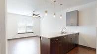 This single-floor unit offers a thoughtfully designed open-concept layout that seamlessly connects the kitchen and living area. The kitchen serves as the focal point, anchored by a peninsula with a granite countertop, an undermount double sink, and rich dark-wood cabinetry below. Above, sleek gray upper cabinets provide ample storage, while a trio of pendant lights and an adjustable track lighting system illuminate the space with style. Stainless steel and black appliances — including a refrigerator, range, and built-in microwave — complete the well-equipped kitchen. The living area extends naturally from the kitchen and features warm hardwood-style flooring and a ceiling fan, creating a comfortable and functional gathering space. A window with horizontal blinds allows natural light to flow in. The bedroom is finished with plush carpeting, a ceiling fan, and a window overlooking the outdoors, offering a quiet and restful retreat. The bathroom is clean and contemporary, featuring large-format gray tile surrounding a soaking tub and shower combination, with dark wood overhead cabinetry for additional storage. A dedicated laundry area with a washer and dryer adds everyday convenience. Throughout the unit, consistent use of gray tones, dark wood accents, and hardwood-style flooring creates a cohesive and modern aesthetic.