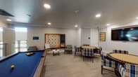 Located at 25500 Westheimer Pkwy in Katy, TX, this dedicated game room is a thoughtfully designed recreational space built for social engagement and leisure. The room features light wood-look flooring, neutral gray walls, and crown molding throughout, creating a clean, modern aesthetic accented by recessed lighting that evenly illuminates the space.

A full-size pool table with a bold blue felt surface serves as a central focal point, providing ample room for play. Adjacent to the pool area, an oversized wall-mounted Scrabble board framed in warm wood offers a standout feature ideal for group wordplay, accompanied by a chalkboard and a whiteboard easel nearby. A dartboard station with a cork surround is mounted on the wall, providing another engaging activity option.

The card and game table area includes two round poker-style tables surrounded by plaid upholstered chairs, creating comfortable seating arrangements for multiple players. A wall-mounted flat-screen TV is positioned to serve this seating area, adding a media component to the room. Decorative backgammon-themed artwork adorns the walls, reinforcing the game-centric theme.

An open railing overlooks an adjacent lower-level area, adding architectural interest and a sense of openness to the space. This game room is well-suited for community or shared-use environments, offering diverse entertainment options in one cohesive, inviting setting.