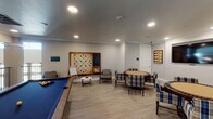 Located at 25500 Westheimer Pkwy in Katy, TX, this dedicated game room is a thoughtfully designed recreational space built for social engagement and leisure. The room features light wood-look flooring, neutral gray walls, and crown molding throughout, creating a clean, modern aesthetic accented by recessed lighting that evenly illuminates the space.

A full-size pool table with a bold blue felt surface serves as a central focal point, providing ample room for play. Adjacent to the pool area, an oversized wall-mounted Scrabble board framed in warm wood offers a standout feature ideal for group wordplay, accompanied by a chalkboard and a whiteboard easel nearby. A dartboard station with a cork surround is mounted on the wall, providing another engaging activity option.

The card and game table area includes two round poker-style tables surrounded by plaid upholstered chairs, creating comfortable seating arrangements for multiple players. A wall-mounted flat-screen TV is positioned to serve this seating area, adding a media component to the room. Decorative backgammon-themed artwork adorns the walls, reinforcing the game-centric theme.

An open railing overlooks an adjacent lower-level area, adding architectural interest and a sense of openness to the space. This game room is well-suited for community or shared-use environments, offering diverse entertainment options in one cohesive, inviting setting.