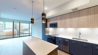 Located at 600 N Glebe Rd in Arlington, Virginia, this modern single-floor residence showcases a thoughtfully designed open layout with contemporary finishes throughout. The kitchen is a standout feature, boasting two-tone cabinetry that combines navy blue lower cabinets with warm wood-toned uppers, crisp white quartz countertops, a large center island, and a full suite of stainless steel appliances including a French door refrigerator, range, microwave, and dishwasher. Stylish pendant lighting in brushed gold and black adds a sophisticated touch overhead. The living area features warm wood-tone laminate flooring and floor-to-ceiling windows that frame city views and open onto a private balcony overlooking N Glebe Road and the Arlington skyline. The bedrooms are finished with plush carpeting and large windows that allow ample natural light while providing views of the surrounding urban landscape. The bathroom is finished in a clean, minimalist style with wood-look flooring, a floating dark-toned vanity, white countertop, and a glass-enclosed walk-in shower with white subway tile. The overall aesthetic is modern and cohesive, blending neutral tones with bold accent colors for a refined urban living experience.