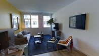 This thoughtfully designed single-floor space exudes a clean, contemporary aesthetic that balances comfort and style. The living area serves as the centerpiece of the space, anchored by large windows that invite abundant natural light and offer views of the surrounding building exterior. The hardwood flooring adds warmth and continuity throughout, complemented by a deep navy area rug that defines the seating zone. A streamlined sofa and a striking gold-framed accent chair with an orange throw provide stylish yet comfortable seating arranged around a geometric coffee table and a compact side table. A flat-screen television sits atop a low-profile media console, flanked by a lush indoor fiddle-leaf fig tree that introduces a natural, organic element to the room. Neutral gray walls create a calm, versatile backdrop, enhanced by carefully selected wall art, including a framed blue-toned abstract piece that adds visual interest. A warm floor lamp in the corner contributes to the layered lighting scheme. The overall ambiance is modern, curated, and welcoming, reflecting an urban sensibility with a well-considered mix of textures, tones, and furnishings that make the space both functional and aesthetically refined.