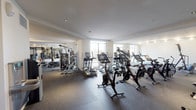 This expansive fitness center is thoughtfully designed to accommodate a full range of workout disciplines across a single open floor. The space is organized into several distinct functional zones, each outfitted with professional-grade equipment. The cardio area features a generous lineup of treadmills, ellipticals, stationary and spin bikes, a rowing machine, and a stair climber, all positioned near large windows that allow natural light to fill the room and provide views to the exterior. A dedicated strength training section houses a multi-station cable machine, selectorized resistance machines targeting various muscle groups, and a power rack with barbells and Olympic plates accompanied by a flat bench. An adjacent free weights zone offers a full dumbbell rack paired with adjustable benches and a large wall-mounted mirror, enhancing both functionality and the sense of space. A supplementary area provides medicine balls, resistance bands, stability balls, and a hydration station for added convenience. Throughout the facility, rubber flooring provides durability and comfort underfoot, while a combination of recessed and track lighting ensures consistent, well-distributed illumination. The clean white walls and contemporary finishes contribute to an efficient, professional atmosphere suited to a wide variety of fitness routines.