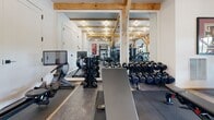 This fitness room combines rustic architectural character with a full suite of professional-grade exercise equipment. Exposed timber beam framing rises toward vaulted ceilings, lending warmth and a distinctly lodge-inspired aesthetic to the space. White walls and recessed lighting keep the interior bright, while multiple windows along one wall invite natural light and views of the surrounding greenery.

The room is organized to accommodate a wide range of workout styles. The cardio zone features dual treadmills, an elliptical, a stair climber, and a spin bike, all positioned to take advantage of the natural light from the windows. A rowing machine with a digital display adds to the cardio offerings.

The strength training area is equally well-appointed, with a full rack of dumbbells, a weight bench, and a multi-functional power rack equipped with a cable system, pull-up bar, and attached weight plates. A heavy punching bag rounds out the functional training options.

Durable rubber and interlocking floor mats cover the workout areas, providing both safety and cushioning. Large mirrors on one wall visually expand the space. The room is accessible via glass-paned double doors, reinforcing its open, welcoming character.