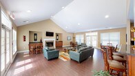 This single-floor community clubhouse is designed to provide a comfortable and functional gathering space with an open, airy layout. The main lounge area immediately impresses with its soaring vaulted ceilings and recessed lighting, creating a bright and expansive atmosphere. Rich hardwood-style flooring flows throughout, anchoring the warm, neutral-toned walls that give the space an inviting character.

The central lounge is furnished with plush leather seating arranged around a glass-top coffee table and a colorful area rug, all oriented toward a classic white-mantled fireplace topped with a wall-mounted flat-screen television. A credenza with dark granite countertops flanks the fireplace, adding to the room's polished aesthetic.

Multiple dining areas are distributed throughout the space, each featuring wooden tables and chairs. Large windows and sets of French doors flood the interior with natural light and provide views of the outdoor pool area, seamlessly connecting the interior to the exterior.

The kitchen is well-appointed with rich cherry wood cabinetry, dark granite countertops, a stainless steel dishwasher, a double sink, and a microwave. A peninsula with bar-height seating extends into the space, making it ideal for casual gatherings or serving guests. The kitchen is finished with neutral tile flooring, contrasting pleasantly with the hardwood of the main lounge.