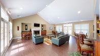 This single-floor community clubhouse is designed to provide a comfortable and functional gathering space with an open, airy layout. The main lounge area immediately impresses with its soaring vaulted ceilings and recessed lighting, creating a bright and expansive atmosphere. Rich hardwood-style flooring flows throughout, anchoring the warm, neutral-toned walls that give the space an inviting character.

The central lounge is furnished with plush leather seating arranged around a glass-top coffee table and a colorful area rug, all oriented toward a classic white-mantled fireplace topped with a wall-mounted flat-screen television. A credenza with dark granite countertops flanks the fireplace, adding to the room's polished aesthetic.

Multiple dining areas are distributed throughout the space, each featuring wooden tables and chairs. Large windows and sets of French doors flood the interior with natural light and provide views of the outdoor pool area, seamlessly connecting the interior to the exterior.

The kitchen is well-appointed with rich cherry wood cabinetry, dark granite countertops, a stainless steel dishwasher, a double sink, and a microwave. A peninsula with bar-height seating extends into the space, making it ideal for casual gatherings or serving guests. The kitchen is finished with neutral tile flooring, contrasting pleasantly with the hardwood of the main lounge.
