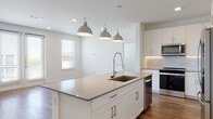This single-floor unit is designed with a clean, contemporary aesthetic that prioritizes open living and functional design. The heart of the home is a spacious open-concept kitchen and living area unified by warm hardwood-style flooring. The kitchen is outfitted with white shaker cabinetry, a marble-style tile backsplash, quartz countertops, and a full suite of stainless steel appliances including a French door refrigerator, range, microwave, and dishwasher. A large center island with an undermount sink and a trio of industrial-style pendant lights serves as both a prep space and a visual anchor for the room. The living area benefits from multiple windows and French doors that invite abundant natural light and offer views to the surrounding neighborhood. Two bathrooms provide distinct configurations: one features a glass-enclosed walk-in shower, LED-backlit vanity mirror, white cabinetry, and quartz countertops; the other offers a full soaking tub with tile surround, an extended double vanity, and matching finishes. Recessed lighting throughout complements the bright, neutral palette, and an in-unit laundry area adds everyday convenience. The overall ambiance is polished, light-filled, and move-in ready.