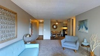 This single-floor residence offers a cohesive and functional layout with a comfortable, mid-century modern aesthetic. The living area serves as the heart of the space, featuring plush carpeting in a warm neutral tone, complemented by cool grey walls that create a calm and inviting atmosphere. A pair of light blue accent chairs — one a classic sofa silhouette and the other a sleek armchair with tapered wooden legs — anchor the seating area, adding a retro-inspired pop of color. A sculptural chrome table lamp and an abstract wall painting introduce artistic character to the space.

The open layout flows seamlessly into a dedicated dining area, where a round wooden dining table and cushioned chairs are centered beneath a ceiling fan and a decorative sunburst mirror, adding visual interest to the wall. Natural light filters through the space, enhancing its airy feel.

Adjacent to the dining area, the kitchen is visible through the open-concept design, featuring dark cabinetry and appliances that provide a grounded contrast to the lighter living spaces. A hallway with a white entry door is visible in the background, offering a clear sense of the home's functional flow and accessibility throughout.