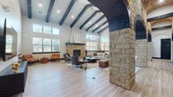 This single-story space makes a striking architectural statement through its bold combination of rustic and contemporary design elements. The main living area commands attention with its soaring vaulted ceiling adorned with dark-stained exposed wood beams set against white-painted shiplap, flooding the room with a sense of grandeur. Large windows positioned at multiple heights draw in abundant natural light, enhancing the open, airy atmosphere throughout.

A defining architectural feature is the series of substantial limestone-clad arched columns and passageways, their warm sandy tones contrasting beautifully with the dark beam accents above. The central living space is anchored by a limestone fireplace with a bold dark mantle, complemented by light wood-look flooring that runs continuously throughout the interior.

A dedicated media area offers wall-mounted display space, while the kitchen is outfitted with dark cabinetry, white quartz countertops, a large center island with bar seating, a farmhouse-style stainless steel sink, open wood shelving, and a full suite of stainless steel appliances. Pendant lighting above the island adds a modern, polished finish. The overall ambiance balances refined rustic character with clean, modern detailing.