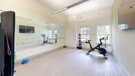 Located at 243 Steele Rd in West Hartford, Connecticut, this dedicated exercise room is a thoughtfully designed fitness space that offers a comprehensive workout environment. The room features light-toned luxury vinyl plank flooring that lends a clean, modern aesthetic throughout.

A full-length wall mirror paired with a classic wooden ballet barre serves as a central feature, making the space highly functional for both dance-based and general fitness training. The room is illuminated by sleek, suspended linear LED light fixtures mounted on high ceilings, contributing to an open and airy atmosphere. Two large double-hung windows along one wall frame views of the surrounding greenery, flooding the space with natural light.

The exercise room is outfitted with a variety of equipment, including a Peloton cycling bike, a tiered medicine ball rack, exercise mats stored on a freestanding rack, and a small trampoline. A wall-mounted flat-screen television is positioned for convenient viewing during workouts. The room also includes a sanitary wipe dispenser, reflecting a commitment to cleanliness and user comfort.

A set of glass-paneled double doors provides direct access to an adjacent corridor, enhancing the sense of connectivity within the broader facility. The overall design of the space balances functionality with a bright, welcoming ambiance suited for a wide range of fitness activities.