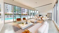 Located at 200 W Ocean Blvd in Long Beach, California, this single-floor amenity space offers a sophisticated, open-plan layout designed for comfort and social gathering. The interior is defined by its clean, contemporary aesthetic, featuring polished concrete floors, crisp white walls, and soaring ceilings that amplify the sense of openness throughout.

Floor-to-ceiling glass walls and sliding doors dominate the perimeter of the space, flooding the interior with abundant natural light and providing sweeping, unobstructed views of the resort-style outdoor pool deck, complete with lounge chairs, umbrellas, and palm trees. The seamless indoor-outdoor connection creates an airy, California-casual atmosphere ideal for both relaxation and entertaining.

The living area is furnished with a plush white sofa, accent chairs, and a low-profile white marble coffee table, all arranged to encourage conversation and poolside views. Adjacent to the lounge area, a generous dining space accommodates a large wooden table surrounded by modern white chairs. The fully integrated kitchenette features sleek floor-to-ceiling white cabinetry, a stainless steel refrigerator, microwave, and sink — providing full functionality within a minimal footprint. Direct access to an outdoor terrace with additional seating further extends the entertaining possibilities of this thoughtfully designed community space.