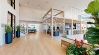 Located at Laurel Sound in Sanford, Florida, this generously proportioned single-floor clubhouse delivers an impressive community gathering destination. The architectural design centers on soaring double-height ceilings and a striking open-plan layout, unified by warm light wood flooring and crisp white walls that create a bright, welcoming atmosphere throughout.

The grand entry foyer sets the tone immediately, featuring floor-to-ceiling black-framed glass doors flanked by tall navy cylindrical planters and a sweeping staircase with natural wood treads and black metal railings. A signature structural pergola of light wood beams defines the central social hub, beneath which pendant globe lights hang above a long communal bar and counter seating with teal-upholstered stools.

Multiple distinct lounge zones offer flexible gathering spaces. One features herringbone-patterned tile flooring, rattan armchairs, and oversized windows framing outdoor views of palm trees and the surrounding community. Another lounge is anchored by a sculptural pendant fireplace and floor-to-ceiling glazing that floods the room with Florida sunshine. A separate, more casual seating area with a teal sofa and woven pendant lights provides additional relaxed comfort.

A dedicated café station with light wood cabinetry, a commercial coffee machine, and open shelving rounds out the amenities, making this a fully equipped and beautifully designed community centerpiece.