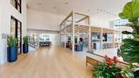 Located at Laurel Sound in Sanford, Florida, this generously proportioned single-floor clubhouse delivers an impressive community gathering destination. The architectural design centers on soaring double-height ceilings and a striking open-plan layout, unified by warm light wood flooring and crisp white walls that create a bright, welcoming atmosphere throughout.

The grand entry foyer sets the tone immediately, featuring floor-to-ceiling black-framed glass doors flanked by tall navy cylindrical planters and a sweeping staircase with natural wood treads and black metal railings. A signature structural pergola of light wood beams defines the central social hub, beneath which pendant globe lights hang above a long communal bar and counter seating with teal-upholstered stools.

Multiple distinct lounge zones offer flexible gathering spaces. One features herringbone-patterned tile flooring, rattan armchairs, and oversized windows framing outdoor views of palm trees and the surrounding community. Another lounge is anchored by a sculptural pendant fireplace and floor-to-ceiling glazing that floods the room with Florida sunshine. A separate, more casual seating area with a teal sofa and woven pendant lights provides additional relaxed comfort.

A dedicated café station with light wood cabinetry, a commercial coffee machine, and open shelving rounds out the amenities, making this a fully equipped and beautifully designed community centerpiece.