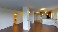 This single-floor residence is designed with an open-concept layout that maximizes the sense of space and flow throughout the living areas. Rich, dark hardwood flooring extends across the main living space, lending warmth and a polished aesthetic to the interior.

A defining architectural feature is the structural white column near the center of the space, which incorporates a small granite-topped ledge, adding both character and a functional surface. The open living area transitions smoothly toward the kitchen, which is positioned along one side and features warm wood cabinetry, granite countertops, and a breakfast bar overhang that invites casual seating. Pendant lighting fixtures are suspended above the kitchen area, providing focused task lighting while enhancing the overall ambiance.

The space is illuminated by a combination of flush-mount ceiling fixtures and pendant lights, giving the interior a warm, inviting glow. A hallway visible from the main living area leads to additional rooms, including a bathroom that is partially visible and features a bright, warmly lit interior with standard fixtures.

White walls throughout keep the space feeling clean and open, while the neutral palette provides a versatile backdrop. The single-floor layout ensures easy navigation, making the space functional and accessible.