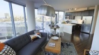 This contemporary single-floor residence showcases a thoughtfully designed open-concept layout that seamlessly integrates the living, dining, and kitchen areas into one cohesive and functional space. Floor-to-ceiling windows wrap around the main living area, flooding the interior with natural light and framing sweeping panoramic city views that serve as a dramatic backdrop throughout.

The living area is anchored by a comfortable sectional sofa, a sleek wooden coffee table, and a textured area rug, all complemented by a striking oversized pendant light that adds a bold design statement. Rich dark hardwood flooring runs throughout, adding warmth and continuity to the space.

The dining area transitions naturally from the living room, featuring a wooden dining table surrounded by clean-lined white chairs — an elegant contrast to the darker tones found elsewhere in the space.

The kitchen is outfitted with dark cabinetry, stainless steel appliances including a built-in microwave and range, and a white kitchen island that provides additional prep and casual seating space. Track lighting above the kitchen ensures excellent task illumination. Together, these elements create a sophisticated urban living environment that balances modern aesthetics with everyday practicality.