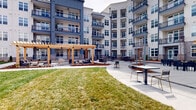 This expansive outdoor courtyard is nestled within a contemporary multi-story residential community, enclosed by buildings featuring a neutral palette of light stucco and slate-blue paneling, large windows, and private balconies. The courtyard is thoughtfully designed to offer a variety of functional zones connected by wide, curving concrete pathways. A central open lawn provides a generous green space, offering a natural focal point surrounded by landscaped beds filled with mulch and young trees.

A prominent wood-framed pergola serves as the centerpiece of the space, providing partial shade over an outdoor lounge area furnished with sectional seating. Adjacent to it, a covered outdoor kitchen features a substantial granite-topped bar built from brick, equipped with professional-grade grills — ideal for communal gatherings. String lights are strung between pergola structures, creating an inviting atmosphere for evening use.

Multiple dining and café-style seating areas with dark metal tables and chairs are distributed throughout the paved sections, accommodating both small groups and larger gatherings. Landscape lighting integrated throughout the grounds enhances the courtyard's usability after dark. The overall design balances open green space with structured social areas, creating a versatile outdoor amenity.