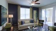 This single-story space in Lewisville, Texas, presents a thoughtfully designed living area that balances bold color choices with contemporary furnishings. The living room serves as the focal point, featuring striking navy blue accent walls that create a dramatic backdrop for the neutral-toned sofa, which is styled with an array of blue and green decorative pillows. A patterned blue area rug anchors the seating area and adds visual texture to the space.

Natural light filters into the room through a large window dressed with white horizontal blinds and floor-length patterned drape panels in a complementary green and cream palette. A ceiling fan with dark wood-toned blades is centrally mounted overhead, providing both airflow and a functional design element. An additional window or sliding glass door on the adjacent wall further enhances the natural light within the room.

Accent pieces include a glass-top coffee table, a woven basket side table, a dark green armchair, and a large framed landscape artwork that ties together the room's green and blue color scheme. The overall ambiance is modern and curated, with a cohesive color story throughout.