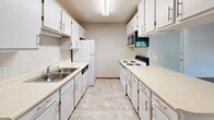 This single-floor unit offers a practical and efficiently arranged layout, accommodating three bedrooms, one bathroom, and a dedicated kitchen within a compact footprint. Neutral gray and beige tones carry throughout the space, contributing to a clean, understated aesthetic.

The living area features plush wall-to-wall carpeting, a textured ceiling, and a broad window that introduces natural light and outdoor views, creating an open and welcoming atmosphere. Adjacent to the living space, the galley-style kitchen is well-equipped with ample white cabinetry on both sides, light countertops, tile flooring, and standard appliances including a refrigerator, electric range, dishwasher, and microwave.

The primary bedroom is a comfortable size, featuring a ceiling fan, a window with blinds, and carpet flooring. The remaining two bedrooms are more compact, each with carpet flooring, windows with blinds, and closets fitted with wire shelving. One bedroom includes a walk-in closet with additional wire rack storage.

The single full bathroom features tile flooring, a combined tub and shower with sliding glass doors, a toilet, and a vanity with built-in cabinetry, maintaining the consistent neutral palette found throughout the unit.