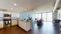 Located at 1167 Front Ave in Cleveland's Flats neighborhood, this single-floor residence showcases a refined industrial-modern aesthetic defined by exposed concrete ceilings, rich dark hardwood flooring, and dramatic floor-to-ceiling windows throughout. The open-concept living and kitchen area forms the heart of the space, where abundant natural light pours in and panoramic views of the surrounding urban landscape and nearby bridge infrastructure create a striking backdrop.

The kitchen is well-appointed with crisp white cabinetry extending to the ceiling, granite countertops, stainless steel appliances, and a large center island with bar seating — ideal for both cooking and casual dining. Pendant lighting adds warmth to the area. The adjacent living room is anchored by the expansive window wall, offering views of the neighborhood and beyond, with access to a private balcony.

The bedroom continues the concrete ceiling detail and features large sliding glass doors opening to the balcony, providing additional natural light and outdoor views. A second bedroom offers a serene retreat with soft accent walls and recessed lighting. The bathroom is finished with a floating vanity, granite countertop, vessel sink, and large-format tile flooring, reflecting the same clean, contemporary design found throughout the entire residence.