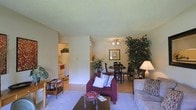 This single-floor residence offers a comfortable and well-organized layout designed for everyday living. The open-concept living and dining area serves as the heart of the home, creating a seamless flow between spaces. The living area features soft carpeting underfoot, a dark wood coffee table, and plush seating, fostering a warm and inviting atmosphere. A large decorative plant adds a natural, lively touch to the corner of the room. Adjacent to the living area, the dining space is finished with hardwood-style flooring and centers around a round dining table with dark chairs, illuminated by a ceiling fan with integrated lighting. Framed wall art and additional greenery add character and warmth to the dining area. A partial wall divides the living space from the dining area, providing definition while maintaining an open feel. The kitchen is visible through a connecting doorway and features white appliances, including a refrigerator and stove, set against light cabinetry. The textured ceilings throughout the space add a classic architectural detail, while the neutral wall tones create a bright and airy ambiance that ties all areas of the home together cohesively.