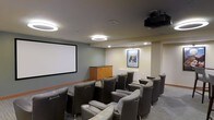 This dedicated screening room delivers a true cinematic experience within a refined, comfortable setting. The space is thoughtfully designed with rows of plush, high-back reclining chairs arranged in a theater-style layout, each equipped with integrated armrests and cup holders for added convenience. Seating is oriented toward a large, wall-mounted projection screen that serves as the room's focal point, complemented by a ceiling-mounted projector that ensures a high-quality viewing experience. The room is illuminated by distinctive circular pendant light fixtures suspended from a smooth, gray ceiling, alongside recessed downlights that provide ambient and accent lighting throughout. The neutral gray walls create a calm, focused atmosphere well-suited for screenings, while warm wood cabinetry near the front of the room offers practical storage. A framed movie poster adds a charming, cinema-inspired decorative touch to the rear wall. Carpeted flooring in a muted tone contributes to the room's acoustic comfort and overall polished aesthetic. This space is ideally suited as a shared amenity, offering residents or guests a private, well-equipped venue for movie nights and group viewings.