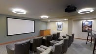 This dedicated screening room delivers a true cinematic experience within a refined, comfortable setting. The space is thoughtfully designed with rows of plush, high-back reclining chairs arranged in a theater-style layout, each equipped with integrated armrests and cup holders for added convenience. Seating is oriented toward a large, wall-mounted projection screen that serves as the room's focal point, complemented by a ceiling-mounted projector that ensures a high-quality viewing experience. The room is illuminated by distinctive circular pendant light fixtures suspended from a smooth, gray ceiling, alongside recessed downlights that provide ambient and accent lighting throughout. The neutral gray walls create a calm, focused atmosphere well-suited for screenings, while warm wood cabinetry near the front of the room offers practical storage. A framed movie poster adds a charming, cinema-inspired decorative touch to the rear wall. Carpeted flooring in a muted tone contributes to the room's acoustic comfort and overall polished aesthetic. This space is ideally suited as a shared amenity, offering residents or guests a private, well-equipped venue for movie nights and group viewings.