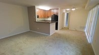 This single-floor residential space offers a practical and open layout designed for comfortable everyday living. The kitchen serves as a central feature, showcasing warm medium-toned wood cabinetry, dark countertops, and black appliances including a full-size refrigerator. A peninsula countertop extends outward, creating a natural divider between the kitchen and the main living area while also providing additional prep and serving space. An overhead fluorescent light fixture illuminates the kitchen workspace efficiently. The open living area is finished with light neutral carpeting that extends throughout, lending a cohesive and inviting feel to the space. The adjoining room, visible through a wide open passage, is bright and airy, benefiting from natural light and a ceiling fan that adds both functionality and a casual aesthetic. The overall color palette features soft, neutral tones — warm beige walls and light flooring — creating a calm and adaptable environment. The open-concept flow between the kitchen, living area, and additional room allows for flexible use of the space and a sense of generous square footage throughout the home.