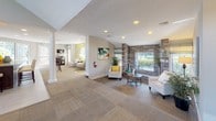 This single-floor community clubhouse offers a thoughtfully designed space that blends comfort and functionality in an inviting, contemporary setting. The open-plan layout flows seamlessly between distinct zones, each styled with coordinated teal, cream, and yellow accents that lend the space a cohesive, upbeat atmosphere.

The main lounge area is anchored by soaring ceilings that rise to accommodate clerestory windows, flooding the room with natural light and offering views of the lush outdoor greenery. Plush sectional seating, a geometric coffee table, and patterned carpet tiles create a relaxed gathering space ideal for socializing or unwinding.

An adjacent seating area features elegant white column accents, vaulted ceilings, and a stone-clad accent wall framing a large window. Comfortable chairs and curated artwork on warm yellow walls give this zone a gallery-like warmth. Decorative plants and framed coastal-inspired artwork are distributed throughout, reinforcing the space's cheerful, resort-style character.

The refreshment station is well-appointed with granite countertops, dark cabinetry, a professional-grade coffee machine, microwave, and under-counter refrigerator — all set beneath bright windows with slatted blinds. Light tile flooring extends through this area toward a glass door that opens to an outdoor patio.

Completing the space is a tidy hallway with dual water fountains and restroom access, accented with colorful framed artwork for a polished, welcoming finish.