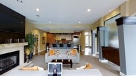 This single-floor space showcases a thoughtfully designed open-concept layout that seamlessly blends the living and kitchen areas into one cohesive environment. The living area is anchored by a pair of plush gray sofas accented with vibrant orange and patterned throw pillows, arranged around a dark wood coffee table atop a light area rug. A striking floor-to-ceiling fireplace with sleek dark framing serves as a dramatic focal point on one side of the room, while a large potted tree adds a natural, organic touch near the entrance. Recessed lighting throughout the ceiling provides even, modern illumination across the space. The kitchen is positioned at the rear of the open floor plan and features warm wood cabinetry with a suspended range hood, a stainless steel refrigerator, a built-in microwave, and a cooktop. A raised breakfast bar with stylish bar stools provides casual seating and defines the kitchen boundary. Warm earth tones — including golden yellows, deep browns, and soft grays — run throughout the space, creating a welcoming and contemporary ambiance. Large windows allow natural light to filter in, enhancing the open and airy feel of the entire floor.