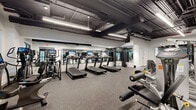 This single-floor fitness center is thoughtfully designed to accommodate a full spectrum of workout needs within a spacious, open-plan layout. The interior features a clean, modern industrial aesthetic, with exposed ductwork and piping set against a dark ceiling, contrasted by crisp white walls and sleek linear LED lighting that ensures bright, even illumination throughout.

The cardio zone is well-appointed with multiple treadmills — each equipped with individual display screens — alongside elliptical trainers and stationary bikes, catering to a variety of cardiovascular training preferences. Large floor-to-ceiling mirrors line the walls, enhancing the sense of space and allowing users to monitor their form during workouts.

The strength training area offers an extensive selection of equipment, including cable machines, adjustable weight benches, squat racks, and a full dumbbell rack stocked with a wide range of weights. Plate-loaded machines and selectorized strength machines provide options for both targeted muscle isolation and compound movements.

Durable rubber flooring covers the entire workout area, providing essential cushioning and traction. Large windows along one wall allow natural light to enter and offer views to the outdoors, adding an airy quality to the environment. The facility is well-organized, ensuring clear circulation paths between equipment zones.