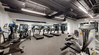 This single-floor fitness center is thoughtfully designed to accommodate a full spectrum of workout needs within a spacious, open-plan layout. The interior features a clean, modern industrial aesthetic, with exposed ductwork and piping set against a dark ceiling, contrasted by crisp white walls and sleek linear LED lighting that ensures bright, even illumination throughout.

The cardio zone is well-appointed with multiple treadmills — each equipped with individual display screens — alongside elliptical trainers and stationary bikes, catering to a variety of cardiovascular training preferences. Large floor-to-ceiling mirrors line the walls, enhancing the sense of space and allowing users to monitor their form during workouts.

The strength training area offers an extensive selection of equipment, including cable machines, adjustable weight benches, squat racks, and a full dumbbell rack stocked with a wide range of weights. Plate-loaded machines and selectorized strength machines provide options for both targeted muscle isolation and compound movements.

Durable rubber flooring covers the entire workout area, providing essential cushioning and traction. Large windows along one wall allow natural light to enter and offer views to the outdoors, adding an airy quality to the environment. The facility is well-organized, ensuring clear circulation paths between equipment zones.