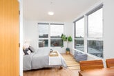 This thoughtfully designed studio unit offers a smart, efficient layout spread across a single floor, combining everyday functionality with a clean, contemporary aesthetic. Light hardwood flooring runs throughout, creating a warm and cohesive feel from entry to living space.

The compact kitchen is positioned near the entrance and is well-equipped with a built-in electric cooktop, an over-the-range microwave, a single-basin sink, and white cabinetry with light countertops. Track lighting overhead ensures the space is bright and practical. The dark appliance finishes provide a modern contrast against the white surfaces.

The open living and sleeping area is the heart of the studio, bathed in abundant natural light pouring through expansive floor-to-ceiling windows. These large windows frame impressive panoramic views of the surrounding urban skyline and neighborhood rooftops, creating a strong sense of connection to the city. The sleeping area is generously proportioned for the overall scale of the unit, with ample wall space and warm, diffused sunlight enhancing the room's calm ambiance.

A dedicated nook near the windows serves as a functional study or lounge area, complete with a wooden desk and seating — ideal for working or simply taking in the views. The overall design of the space emphasizes clean lines, neutral tones, and an airy, open atmosphere throughout.