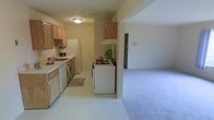 This single-floor residence offers a practical and welcoming layout with a seamless flow between the kitchen and living areas. The kitchen is equipped with light wood cabinetry that provides ample storage space, white appliances including a refrigerator, dishwasher, and a freestanding range, and a countertop workspace ideal for meal preparation. A double-basin sink is positioned beneath upper cabinets, maximizing utility in the kitchen space. The flooring in the kitchen features light-toned tile that transitions smoothly into the carpeted living area. The living space is open and airy, featuring neutral wall tones and carpet flooring that contribute to a warm and comfortable atmosphere. The open-concept design between the kitchen and living room creates a connected, functional environment well-suited for everyday living. Ceiling-mounted lighting provides consistent illumination throughout the space. A doorway visible from the living area suggests additional rooms beyond, hinting at further living or sleeping quarters. Overall, the space presents a clean, understated aesthetic with a focus on functionality and ease of movement throughout the single-level layout.