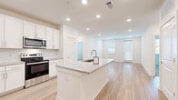This single-floor residence offers a clean, contemporary design with a bright, open layout. The heart of the home is a well-appointed kitchen featuring a large central island topped with granite countertops and an integrated sink. White shaker-style cabinetry extends to the ceiling, providing ample storage, while stainless steel appliances — including a refrigerator with ice maker, electric range, microwave, and dishwasher — complete the space. Recessed lighting illuminates the kitchen and flows seamlessly into the open living area, which is bathed in natural light from multiple windows and offers access to an exterior door. Light hardwood-style flooring runs throughout the main living areas, lending a cohesive and airy feel to the space. The bedroom features soft carpeting and direct access to a bathroom equipped with a soaking tub and tile surround. A walk-in closet with wire shelving provides generous storage. A hallway connects the private areas of the home, with a window at the end drawing in additional natural light. The overall aesthetic is modern and move-in ready, characterized by a neutral white and light wood palette that creates a fresh, inviting atmosphere throughout.