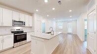 This single-floor residence offers a clean, contemporary design with a bright, open layout. The heart of the home is a well-appointed kitchen featuring a large central island topped with granite countertops and an integrated sink. White shaker-style cabinetry extends to the ceiling, providing ample storage, while stainless steel appliances — including a refrigerator with ice maker, electric range, microwave, and dishwasher — complete the space. Recessed lighting illuminates the kitchen and flows seamlessly into the open living area, which is bathed in natural light from multiple windows and offers access to an exterior door. Light hardwood-style flooring runs throughout the main living areas, lending a cohesive and airy feel to the space. The bedroom features soft carpeting and direct access to a bathroom equipped with a soaking tub and tile surround. A walk-in closet with wire shelving provides generous storage. A hallway connects the private areas of the home, with a window at the end drawing in additional natural light. The overall aesthetic is modern and move-in ready, characterized by a neutral white and light wood palette that creates a fresh, inviting atmosphere throughout.