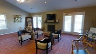This single-floor space functions as a well-appointed communal lounge, thoughtfully designed to provide both comfort and functionality. The centerpiece of the room is a classic tiled fireplace framed by a mantel, above which a wall-mounted flat-screen television is positioned for easy viewing from multiple seating areas. The lounge is furnished with a curated mix of dark-framed armchairs with white cushioned upholstery, arranged in conversational clusters around rustic wooden stump side tables, lending the space a relaxed yet stylish character. The flooring features a warm, multi-toned patterned carpet in rich red and earthy hues, adding texture and color to the interior. Recessed lighting in the ceiling complements the abundant natural light that pours in through multiple white-framed French doors and windows fitted with plantation shutters, creating a bright and airy atmosphere. A large decorative mirror on one wall enhances the sense of space, while informational materials displayed on the mantel suggest this area may serve as a welcoming gathering or reception space. The neutral taupe wall tones tie the design elements together cohesively, resulting in an inviting environment suited for relaxation and social interaction.