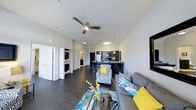 Located at 2131 N. Meridian, this single-floor unit offers an open-concept living space defined by clean lines and modern finishes. The living area features dark hardwood-style flooring that extends throughout, creating a sleek and cohesive aesthetic. A ceiling fan overhead provides both function and a contemporary accent to the bright white ceiling.

The living room is furnished with a spacious sectional sofa and accent chairs in a bold black-and-white pattern, complemented by pops of yellow and teal that add vibrant personality to the space. A glass coffee table anchors the seating arrangement, enhancing the sense of openness.

The kitchen is visible from the living area and features dark cabinetry, black appliances, and a breakfast bar with seating — ideal for casual dining or entertaining. Pendant lighting above the bar adds a refined touch.

To one side, a stacked washer and dryer unit is conveniently integrated into the layout, adding practical value. A hallway leads toward additional living spaces, including what appears to be a bathroom and bedroom. The overall ambiance is modern, energetic, and well-appointed.