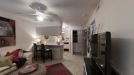 This single-floor residence features an open-concept layout that connects the living and kitchen areas into one cohesive, functional space. The living area is anchored by carpeted flooring, a ceiling fan with integrated lighting, and a textured ceiling, contributing to a cozy, casual ambiance. A large flat-screen television is positioned along one wall, complemented by decorative accents including a tall vase with flowering branches that add a touch of natural elegance.

The kitchen is efficiently designed with white cabinetry, a white refrigerator, microwave, and additional appliances neatly arranged along the walls. A peninsula-style breakfast bar with seating for two serves as both a dining space and a visual divider between the kitchen and living area. Light-toned countertops and tile flooring in the kitchen contrast with the carpeted living zone, clearly delineating each functional area.

A hallway visible toward the rear of the space leads to additional rooms, with a storage closet or pantry featuring shelved interiors. The overall aesthetic is practical and comfortable, with warm, ambient lighting enhancing the welcoming character of the interior.