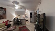 This single-floor residence features an open-concept layout that connects the living and kitchen areas into one cohesive, functional space. The living area is anchored by carpeted flooring, a ceiling fan with integrated lighting, and a textured ceiling, contributing to a cozy, casual ambiance. A large flat-screen television is positioned along one wall, complemented by decorative accents including a tall vase with flowering branches that add a touch of natural elegance.

The kitchen is efficiently designed with white cabinetry, a white refrigerator, microwave, and additional appliances neatly arranged along the walls. A peninsula-style breakfast bar with seating for two serves as both a dining space and a visual divider between the kitchen and living area. Light-toned countertops and tile flooring in the kitchen contrast with the carpeted living zone, clearly delineating each functional area.

A hallway visible toward the rear of the space leads to additional rooms, with a storage closet or pantry featuring shelved interiors. The overall aesthetic is practical and comfortable, with warm, ambient lighting enhancing the welcoming character of the interior.