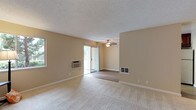 This single-floor residence offers a thoughtfully designed layout across several functional spaces. The living area is generously sized, featuring neutral beige walls, plush carpeting, and a textured ceiling. A ceiling fan with a light fixture anchors the dining or sitting area, while a wall-mounted air conditioning unit and a floor-level heating vent provide climate control year-round. A sliding glass door opens to an outdoor area, allowing natural light to flow in alongside a side window that further brightens the room.

The kitchen is designed for efficiency, featuring rich dark-red cabinetry paired with granite-style countertops that run along both sides of the galley-style layout. Stainless steel appliances, including a dishwasher, refrigerator, and range, are integrated seamlessly, while a skylight-style overhead fixture ensures excellent illumination.

The bedrooms are carpeted and bathed in natural light from windows, with one room offering a wide sliding double-door closet. The bathrooms are finished with matching dark-red vanity cabinetry topped with granite countertops. One bathroom features a tub-shower combination, while the other includes a standalone walk-in shower with glass doors and wood-look flooring, lending a cohesive and polished aesthetic throughout.