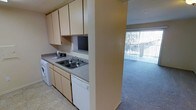 This single-floor residence offers a practical and well-organized layout designed for comfortable everyday living. The kitchen is efficiently arranged along one wall, featuring light wood cabinetry that extends to the ceiling for generous storage capacity. A double-basin sink with a standard faucet is set into a laminate countertop, providing ample workspace for meal preparation. A built-in dishwasher and an in-unit washing machine are seamlessly integrated beneath the counter, adding notable convenience. The kitchen is finished with light tile flooring, offering easy maintenance. A textured dividing wall separates the kitchen from the adjacent living area, creating a sense of definition between the two spaces without fully closing them off. The living area features wall-to-wall carpeting that lends warmth underfoot. A large sliding glass door dressed with vertical blinds allows natural light to fill the space and provides direct access to an outdoor area, offering views of the exterior surroundings. The overall aesthetic is clean and neutral, with warm beige tones throughout that create a cohesive and inviting atmosphere across the entire floor plan.