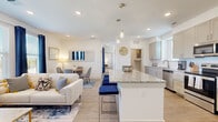 This thoughtfully designed two-story home offers a bright and functional layout across two floors. The lower level houses an attached garage with built-in overhead storage, while the expansive upper floor contains the primary living spaces. At the heart of the home is an open-concept living, dining, and kitchen area bathed in natural light from large windows dressed with navy curtains. The kitchen is a standout feature, showcasing granite countertops, a large center island with bar seating, gray shaker-style cabinetry, a white subway tile backsplash, and a full suite of stainless steel appliances. Light wood-style flooring flows throughout the main living areas, enhancing the airy, modern aesthetic. The dedicated office features a built-in granite-topped L-shaped desk, providing a practical workspace. The spacious primary bedroom is carpeted, filled with natural light, and accompanied by a generous walk-in closet with built-in shelving and hanging rods. The primary bathroom features a dual-sink vanity with granite countertops, gray cabinetry, and hardwood-style flooring. Two additional bedrooms and a utility room round out the home, offering versatile living options throughout.