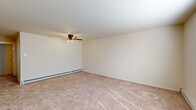 This single-floor, 2-bedroom, 1-bathroom residence offers a straightforward and functional layout across a compact but well-organized footprint. The living room is carpeted and features a ceiling fan with light fixture, a wall-mounted air conditioning unit, and a large window fitted with horizontal blinds that allows natural light to fill the space. A pass-through opening connects the living area to the kitchen, enhancing the sense of openness between the two spaces. The kitchen is equipped with light wood cabinetry, dark countertops, a stainless steel refrigerator, a gas range with an over-the-range microwave, and a single-basin sink. Light tile flooring runs throughout the kitchen. The primary bedroom is generously proportioned and includes a ceiling fan, a walk-in closet with built-in shelving, a window, baseboard heating, and a wall air conditioning unit. The second bedroom, more intimate in scale, is carpeted with neutral walls and offers access to a closet. A notable feature is the in-unit stacked washer and dryer, tucked behind louvered doors in one of the bedrooms. Baseboard heating is present throughout the residence, and the overall palette of neutral tones and consistent carpet creates a cohesive, understated ambiance.