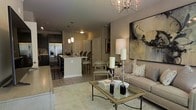 This single-floor residence showcases an open-concept design that integrates the living, dining, and kitchen spaces into one cohesive, flowing layout. The living area is anchored by a neutral-toned sofa paired with a glass-top coffee table featuring wrought iron detailing, all resting on a patterned area rug. A sputnik-style chandelier overhead adds a modern focal point, while a large abstract canvas artwork lends artistic character to the main wall. Tall candle holders and decorative accents contribute to a refined, curated ambiance. The kitchen features dark cabinetry contrasted by light countertops on a peninsula with bar seating, pendant lighting above, and stainless steel appliances including a French door refrigerator. Light hardwood-style flooring runs throughout, unifying the open space. A dining area with upholstered seating bridges the kitchen and living zones, enhancing the sense of connectivity. The overall palette of warm grays and neutral tones, combined with carefully chosen lighting and décor, creates an inviting and elegantly appointed interior.