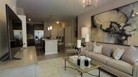 This single-floor residence showcases an open-concept design that integrates the living, dining, and kitchen spaces into one cohesive, flowing layout. The living area is anchored by a neutral-toned sofa paired with a glass-top coffee table featuring wrought iron detailing, all resting on a patterned area rug. A sputnik-style chandelier overhead adds a modern focal point, while a large abstract canvas artwork lends artistic character to the main wall. Tall candle holders and decorative accents contribute to a refined, curated ambiance. The kitchen features dark cabinetry contrasted by light countertops on a peninsula with bar seating, pendant lighting above, and stainless steel appliances including a French door refrigerator. Light hardwood-style flooring runs throughout, unifying the open space. A dining area with upholstered seating bridges the kitchen and living zones, enhancing the sense of connectivity. The overall palette of warm grays and neutral tones, combined with carefully chosen lighting and décor, creates an inviting and elegantly appointed interior.