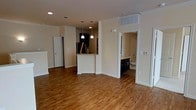 This single-floor space showcases a thoughtfully designed open-concept layout that promotes a sense of spaciousness and connectivity throughout. Warm-toned hardwood flooring extends across the main living area, adding a welcoming and polished aesthetic to the interior.

The kitchen serves as a central focal point, featuring dark cabinetry, a granite or stone-topped peninsula that doubles as a breakfast bar, and pendant lighting suspended above. A stainless steel refrigerator anchors the space, complementing the rich, contrasting tones of the cabinetry. Recessed lighting throughout the ceiling ensures consistent, even illumination across the open floor plan.

Adjacent to the main living area, a bathroom is visible, outfitted with dark vanity cabinetry that mirrors the kitchen's design language, offering a cohesive aesthetic throughout the unit. The bathroom includes a toilet and what appears to be a bathtub, providing full functionality.

Crisp white walls and white-trimmed doorways frame the space cleanly, contributing to a bright and neutral palette that allows for versatile interior styling. The layout flows naturally between the kitchen, living area, and adjoining rooms, making this a functional and visually cohesive single-floor residence.