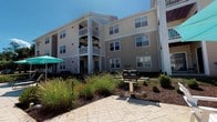 Located at 21265 Mayfaire Ln in Lexington Park, Maryland, this community amenity space at Park Villas offers residents a well-rounded environment for both fitness and outdoor relaxation. The fitness center is thoughtfully equipped with a variety of strength and cardio machines, a cable station, dumbbells, kettlebells, stability balls, and wall-mounted mirrors that enhance the sense of space. Recessed lighting illuminates the carpeted workout area, while windows allow in natural light. A dedicated utility area adjacent to the gym includes a water cooler, a medical-grade scale, and restroom access, ensuring convenience for users. A community map is displayed on the wall, reinforcing the neighborhood identity. Outside, a generously sized stamped concrete patio serves as the heart of the outdoor amenity area. A substantial stone fire pit with built-in seating serves as a central gathering point, surrounded by white Adirondack chairs and teal market umbrellas for shade. Lush ornamental grasses and flowering crape myrtles add natural beauty and color to the landscaped surroundings. A stainless steel gas grill provides outdoor cooking capabilities, making this space ideal for community gatherings. The multi-story residential buildings with white balconies frame the courtyard, creating an inviting and cohesive community atmosphere beneath open blue skies.