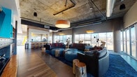 Located at 1420 Oak St in Lakewood, Colorado, this sophisticated single-floor amenity lounge delivers a compelling blend of industrial and contemporary design. The space is defined by its exposed concrete ceilings with visible ductwork and structural elements, contrasted warmly by rich hardwood-style flooring and carefully curated furnishings throughout.

The main lounge area features an expansive open layout anchored by curved leather sectional sofas, tufted ottoman seating in teal, and natural live-edge wood stump accent tables, all set atop a bold, deep-blue patterned area rug. Pendant drum and cube-style light fixtures provide warm, ambient illumination that complements the industrial aesthetic. Floor-to-ceiling windows wrap the perimeter, flooding the space with natural light and offering sweeping views of the surrounding Lakewood landscape and Colorado's Rocky Mountain foothills beyond.

A dedicated kitchen and dining area features a large dark-finished island with bar seating, a warm slatted wood ceiling that transitions the atmosphere from industrial to refined, and an integrated banquette dining nook with wall-mounted artwork. Stainless steel appliances and full-length windows complete the space, keeping the mountain views central even while dining.

A bar counter area with high stools serves as an additional gathering spot, while accent walls in turquoise and bold red add vibrant pops of color throughout, reinforcing the space's energetic and modern character.