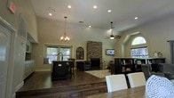 This single-story home showcases an expansive open-concept layout that connects the dining, kitchen, and living areas into one cohesive, flowing space. The vaulted ceilings rise dramatically overhead, enhancing the sense of volume and airiness throughout. Recessed lighting is distributed across the ceiling, supplemented by a classic chandelier over the dining area and a ceiling fan positioned above the living space, ensuring both style and comfort.

The kitchen features a dark-finished island with bar seating, offering a natural transition point between the dining and living zones. Rich hardwood flooring extends throughout, adding warmth and continuity to the open floor plan. The living area is anchored by a floor-to-ceiling stacked stone fireplace, which serves as a striking focal point, with a mounted flat-screen television above. An arched decorative wall accent adds an artistic touch nearby.

Natural light filters in through multiple windows, including an arched window that adds architectural character. The neutral wall tones complement the dark wood and stone finishes, creating a warm, inviting atmosphere well-suited for both everyday living and entertaining.