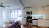 This single-floor space showcases a thoughtfully designed open-concept layout that seamlessly blends the kitchen and living areas into one cohesive environment. The kitchen is outfitted with high-gloss white cabinetry extending to the ceiling, offering generous storage while contributing to a clean, contemporary aesthetic. Stainless steel appliances — including a range with oven, an over-the-range microwave, and a dishwasher — provide a polished, functional cooking space. A white glass backsplash ties the kitchen together with a crisp, modern feel, and ample counter space makes meal preparation comfortable and practical. Recessed lighting throughout the ceiling ensures consistent, even illumination across the space. The living area flows naturally from the kitchen, anchored by rich hardwood flooring that adds warmth and contrast to the otherwise light palette. Large windows invite an abundance of natural light and provide views of the surrounding greenery, creating a bright and airy atmosphere. The overall design balances minimalist sensibility with functional detail, making this a versatile and inviting space well-suited for modern everyday living.